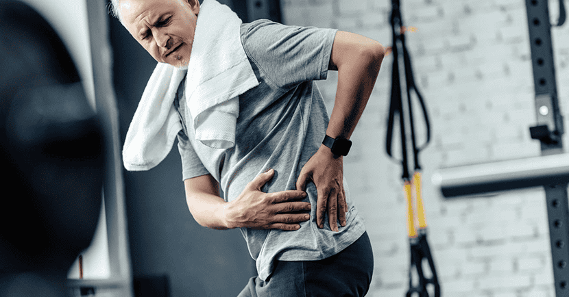 An older man in a gym holding his lower back and side, with a pained expression
