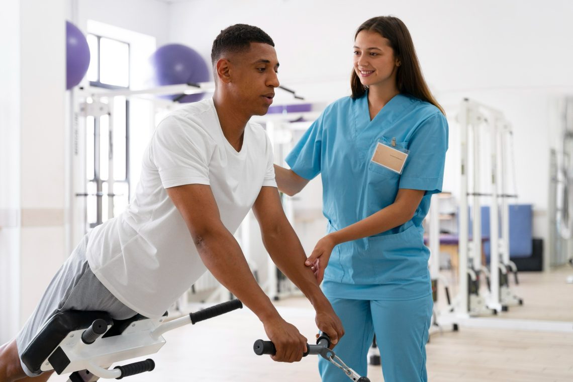 A clinician in scrubs supporting a patient through a seated strengthening exercise in a rehabilitation gym.