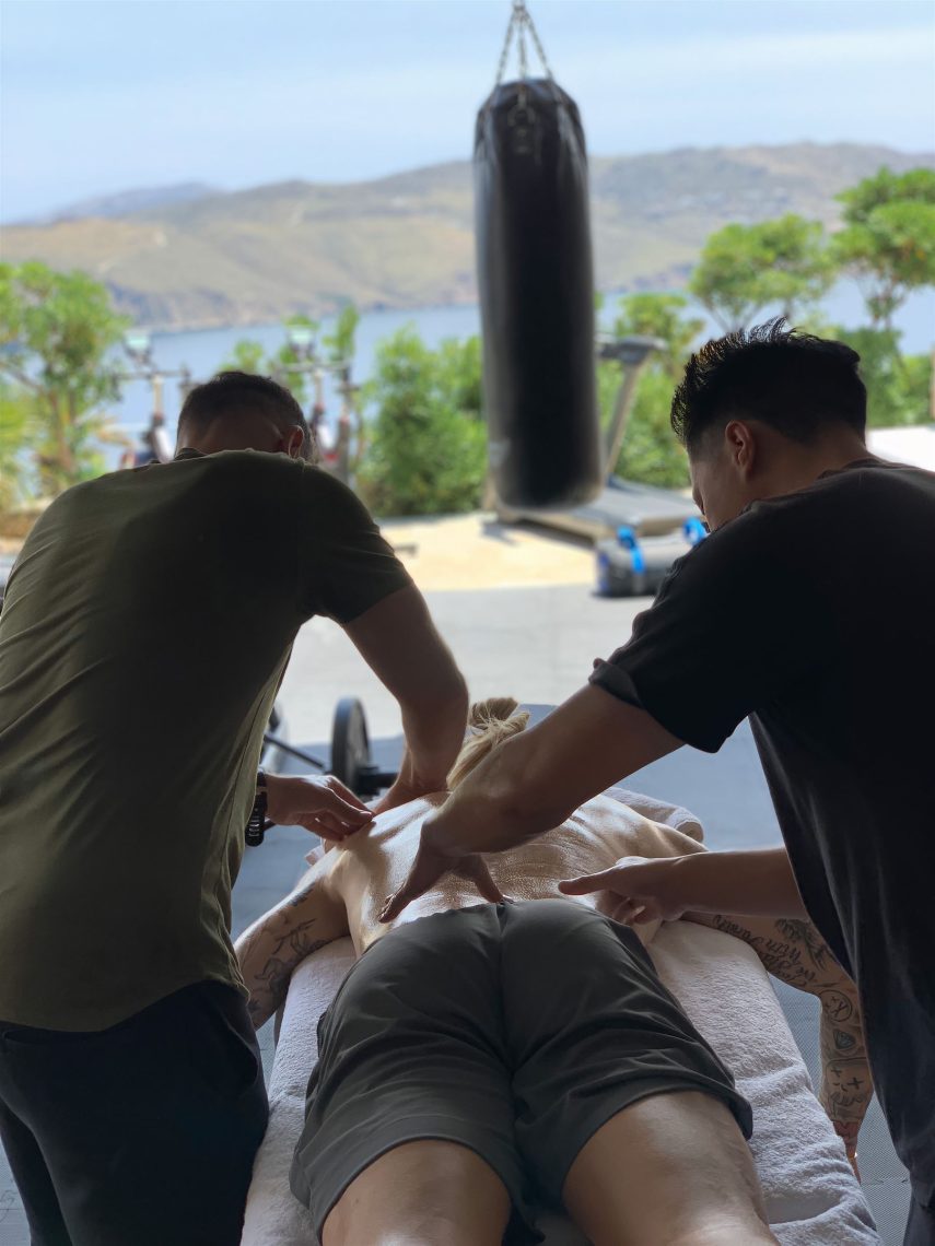 Two therapists treating an athlete lying face down at an outdoor training facility.