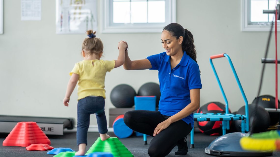 A physiotherapist kneeling to high-five a toddler in a paediatric therapy gym.