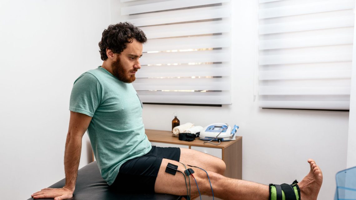 A patient seated on a treatment couch with electrode pads and leads attached to the thigh, beside an electrotherapy unit.