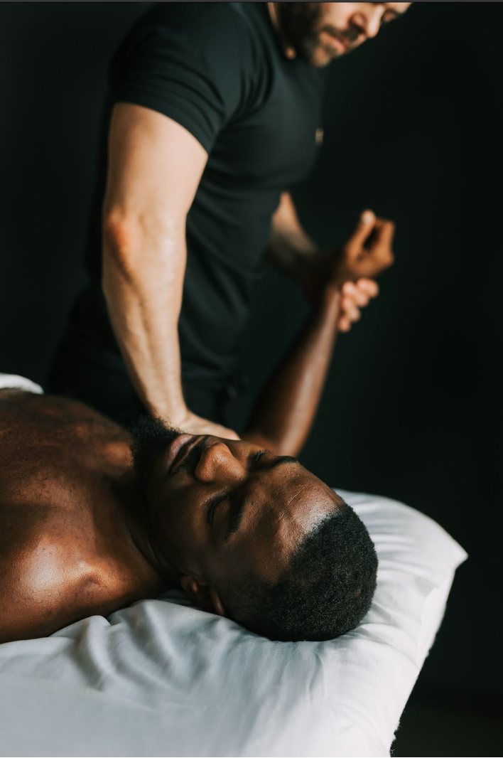 A therapist in a black tee applying a manual therapy technique to the neck and shoulder of a client lying face up on a white-sheeted couch in a dark room.